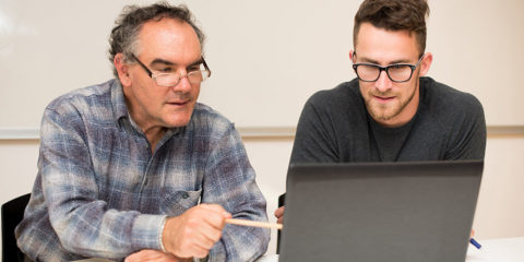 An older male sitting behind a laptop computer, with a younger male. Both are looking at the laptop screen.