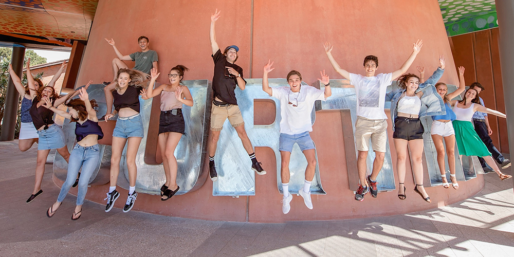 Students leaping in air and smiling in front of Curtin cookie cutter sculpture - play video' }}