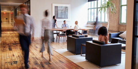 A modern office environment, people walking down a corridor, while others sit on leather couches