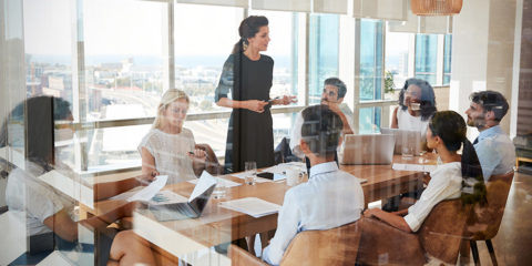 A group of people sitting around a conference table, talking. Image taken through a glass door.